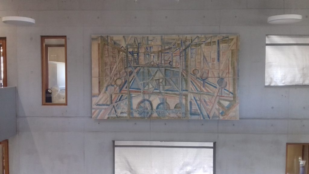 Ein Wandgemälde, das an einer Betonwand hängt. Das Gemälde zeigt eine abstrakte Darstellung von Figuren und Linien in sanften Farben, umgeben von großen Fenstern, die Licht in den Raum lassen.