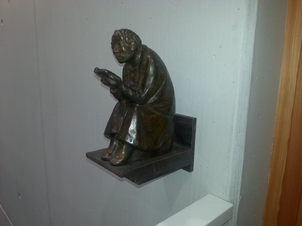 Eine kleine bronzene Skulptur eines sitzenden Mannes, der einen Gegenstand in der Hand hält. Die Figur ist auf einer Wand montiert und zeigt den Mann in einer nachdenklichen Pose.