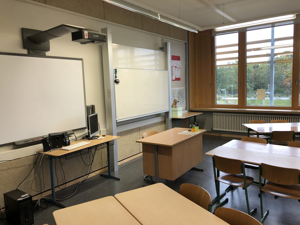 Ein Klassenzimmer mit einem interaktiven Whiteboard, einem Tisch und Stühlen. An der Wand sind Fenster, durch die Tageslicht fällt. Auf dem Tisch steht ein kleiner Behälter. Im Hintergrund sind technische Geräte sichtbar.