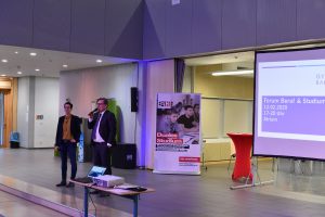 Zwei Personen stehen auf einer Bühne und sprechen in ein Mikrofon. Im Hintergrund ist ein Präsentationsbildschirm zu sehen, auf dem Informationen angezeigt werden. Links steht ein Roll-Up-Banner mit Informationen zu einem Thema, das mit Bildung oder Beratung zu tun hat. Der Raum hat eine moderne Atmosphäre mit gedämpfter Beleuchtung.