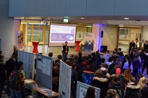 Eine Person steht auf einer Bühne und spricht vor einer großen Gruppe von Zuhörern in einem Innenraum. Im Hintergrund ist ein Präsentationsbildschirm mit Informationen zu sehen. Die Zuhörer sind in einem offenen Bereich versammelt, umgeben von Informationsständen. Der Raum ist hell erleuchtet und hat eine freundliche Atmosphäre.
