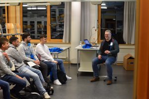 Ein Mann sitzt auf einem Stuhl und spricht vor einer Gruppe von fünf jungen Menschen, die ihm aufmerksam zuhören. Die Szene findet in einem Raum mit großen Fenstern und einem Tisch statt, auf dem ein Laptop und einige Gegenstände liegen. Im Hintergrund sind weitere Stühle und eine Kiste zu sehen. Die Atmosphäre wirkt informell und lehrreich.