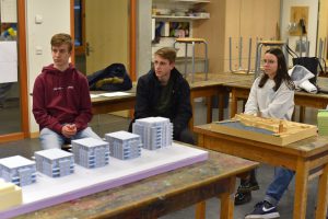 Drei Jugendliche sitzen an einem Tisch in einem Klassenraum. Auf dem Tisch befinden sich mehrere Architekturmodelle, darunter mehrere kleine Gebäude. Im Hintergrund sind weitere Materialien und Möbel zu sehen. Die Jugendlichen schauen aufmerksam in die Richtung der Modelle.
