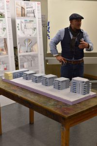 Ein Mann in einem Weste und einer Schirmmütze steht an einem Tisch und erklärt ein Architekturmodell, das mehrere Gebäude darstellt. Im Hintergrund sind Plakate mit Entwürfen und Illustrationen zu sehen. Der Tisch ist aus Holz und hat eine einfache Oberfläche.