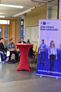 Eine Gruppe von drei jungen Menschen sitzt an einem Tisch in einem Innenraum. Neben ihnen steht ein Stehtisch mit rotem Tischdecken. Im Hintergrund ist ein Werbebanner mit dem Text 