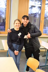 Zwei junge Menschen stehen in einem Klassenzimmer. Die Frau sitzt auf einem Tisch und lächelt, während der Mann neben ihr steht und mit einer Hand ein Zeichen gibt. Im Hintergrund sind Fenster und Tischreihen zu sehen.