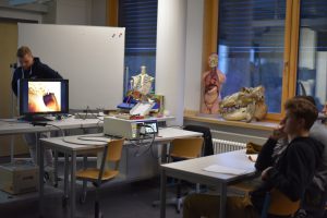 Ein Klassenzimmer mit einem Präsentationsbildschirm, auf dem eine medizinische Grafik zu sehen ist. Im Vordergrund sitzen zwei Schüler an Tischen. Hinter ihnen befinden sich anatomische Modelle, darunter ein Skelett und ein menschliches Modell. Ein Lehrer steht neben dem Bildschirm und erklärt etwas. Der Raum hat große Fenster mit natürlichem Licht.
