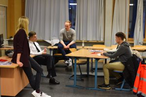 Fünf Personen sitzen und stehen in einem Raum mit Tischen und Stühlen. Eine Person steht an einem Tisch, während die anderen um den Tisch herum sitzen. Die Umgebung wirkt freundlich und informell, mit Vorhängen an den Fenstern.