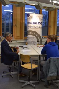 Eine Person sitzt an einem Tisch und spricht mit zwei anderen Personen. Im Hintergrund steht ein Banner mit der Aufschrift 