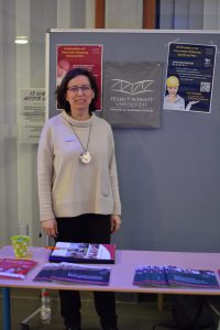 Eine Frau steht an einem Tisch, auf dem mehrere Broschüren und Informationsmaterialien liegen. Im Hintergrund befindet sich ein Plakat mit dem Logo einer Institution. Die Frau trägt einen hellen Pullover und eine auffällige Halskette. Sie schaut freundlich in die Kamera.