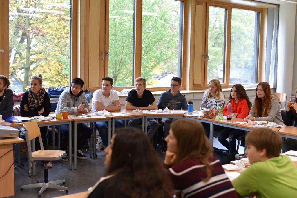 Eine Gruppe von Jugendlichen sitzt an einem großen Tisch in einem hellen Raum mit Fenster, das einen Blick ins Freie bietet. Einige der Teilnehmer hören aufmerksam zu, während andere Notizen machen oder sich unterhalten. Auf dem Tisch liegen verschiedene Getränke und Snacks.