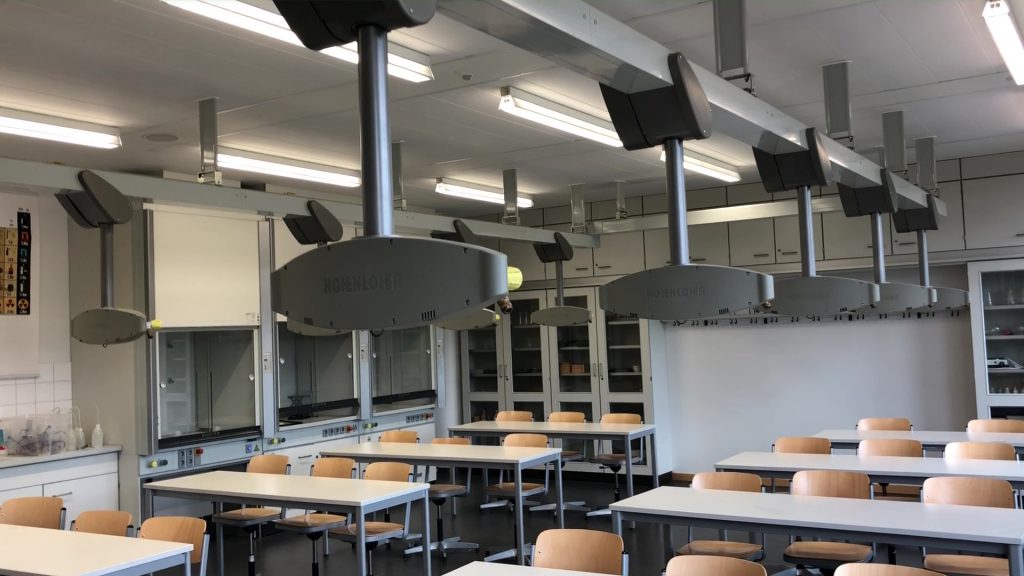 Ein moderner Klassenraum mit mehreren Tischen und Stühlen. An der Decke sind mehrere Belüftungs- oder Abzugshauben installiert. Im Hintergrund sind Regale mit Glasbehältern und anderen Materialien sichtbar. Das Licht ist hell und gleichmäßig.
