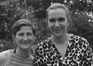 Zwei Frauen lächeln in die Kamera, umgeben von grüner Natur. Die linke Frau trägt ein gestreiftes Oberteil, die rechte trägt ein Oberteil mit einem auffälligen Muster.