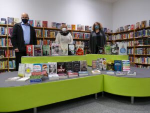 Drei Personen stehen in einer Bibliothek vor einer grünen Bücherinsel, die mit verschiedenen Buchcovern dekoriert ist. Im Hintergrund sind Bücherregale mit vielen Büchern zu sehen. Alle Personen tragen Masken.