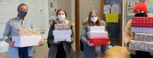 Vier Personen stehen in einem Raum und halten jeweils mehrere Geschenkpäckchen in den Händen. Alle tragen Mund-Nasen-Schutzmasken. Zwei der Personen haben eine graue und eine schwarze Jacke an, die dritte Person trägt einen grünen Mantel und die vierte einen roten Hut. Im Hintergrund sind verschiedene Gegenstände und Plakate sichtbar.