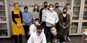 Gruppe von zwölf Personen in einem Raum, einige tragen Schutzmasken. Zwei Personen sitzen im Vordergrund, während die anderen im Hintergrund stehen. Eine Person hält eine Box in der Mitte der Gruppe. Die Umgebung hat Regale mit verschiedenen Gegenständen.