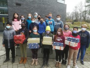 Eine Gruppe von Jugendlichen steht vor einem Gebäude. Sie tragen alle Mund-Nasen-Bedeckungen und halten Geschenkpakete in den Händen. Die Jugendlichen sind in zwei Reihen aufgestellt und lächeln in die Kamera. Im Hintergrund ist ein moderner, grauer Gebäudewall und einige Bäume zu sehen.