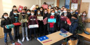 Gruppe von Schülerinnen und Schülern in einem Klassenzimmer, die Masken tragen und Geschenke in den Händen halten. Im Hintergrund ist ein Tisch mit Schulmaterialien zu sehen.