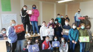 Eine Gruppe von Kindern und Jugendlichen steht in einem Klassenraum und hält Geschenkpäckchen in den Händen. Einige Schüler stehen auf Tischen, während andere auf dem Boden stehen. Alle tragen Mund-Nasen-Schutzmasken. Im Hintergrund sind weitere Schüler und eine Tafel sichtbar. Die Stimmung wirkt fröhlich und gemeinschaftlich.