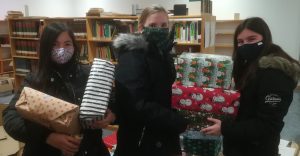 Drei Personen stehen in einem Raum mit Bücherregalen im Hintergrund. Sie tragen Winterjacken und Masken und halten eingepackte Geschenke in den Händen. Zwei Geschenke sind in buntem Geschenkpapier, während eines gestreift ist. Die Atmosphäre wirkt festlich und freundlich.