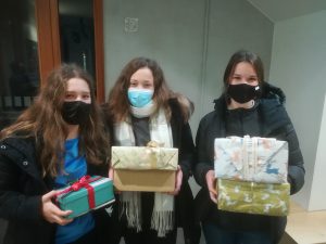 Drei Personen stehen in einem Raum und halten jeweils ein oder mehrere Geschenke in den Händen. Alle tragen Masken. Die Person auf der linken Seite hat ein Geschenk in einer blauen Verpackung mit rotem Band, die mittlere Person hält ein Geschenk in einer neutralen Verpackung, und die Person auf der rechten Seite hält ein Geschenk in einer grünen und weißen Verpackung. Im Hintergrund sind Wände und Fenster zu sehen.