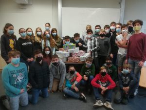 Eine Gruppe von Schülern steht in einem Klassenraum, alle tragen Mund-Nasen-Schutz. Im Hintergrund ist ein Tisch mit mehreren Geschenken zu sehen. Die Schüler posieren zusammen für das Foto, einige lächeln, während andere eine Daumen-hoch-Geste machen.