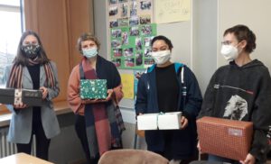 Vier Personen stehen in einem Raum und halten jeweils ein Geschenk in den Händen. Alle tragen Mund-Nasen-Schutzmasken. Im Hintergrund sind einige Bilder an der Wand zu sehen.