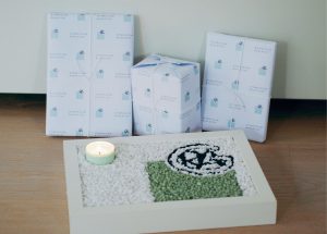 Ein dekoratives Arrangement auf einem Holzboden. Im Vordergrund befindet sich eine flache Box, gefüllt mit weißen und grünen kleinen Steinen, die ein Muster bilden. In der Mitte dieser Box steht eine kleine Kerze. Im Hintergrund sind drei Geschenke, die in weißem Papier mit einem dezenten Muster eingewickelt sind.