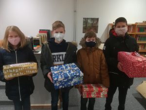 Vier Kinder stehen in einem Raum und halten jeweils ein Geschenk in den Händen. Sie tragen Gesichtsmasken und sind in Winterkleidung. Im Hintergrund sind Regale mit Büchern zu sehen.