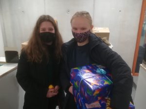 Zwei junge Mädchen stehen in einem Raum mit grauen Wänden. Beide tragen Masken und lächeln. Eines der Mädchen hält ein bunt verpacktes Geschenk in den Händen, während das andere eine gelbe Kugel hält. Im Hintergrund sind Kisten und Möbel zu erkennen.