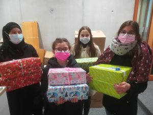 Vier Personen stehen in einem Raum und halten bunt verpackte Geschenke in den Händen. Alle tragen Masken. Im Hintergrund sind Kisten und Möbel zu sehen.