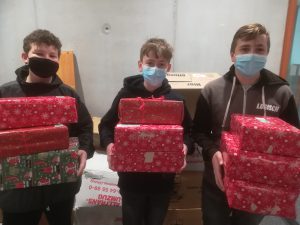 Drei Jugendliche stehen nebeneinander und halten sorgfältig verpackte Geschenke in den Händen. Alle tragen Mund-Nasen-Schutzmasken. Im Hintergrund sind weitere Geschenke und Kartons sichtbar. Die Geschenke sind in festlichem Papier mit roten und grünen Motiven verpackt.