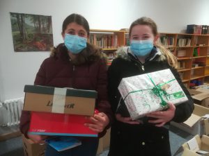 Zwei Jugendliche tragen jeweils ein Geschenk und eine Schachtel. Beide tragen Gesichtsmasken und stehen in einem Raum mit Bücherregalen im Hintergrund.