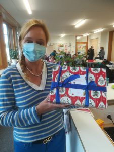 Eine Person mit blonden Haaren, die eine blaue Maske trägt, hält ein bunt verpacktes Geschenk in den Händen. Das Geschenk hat ein rot-weißes Motiv mit weihnachtlichen Elementen und ist mit einer blauen Schleife verziert. Im Hintergrund sind weitere Personen und Büromöbel sichtbar.