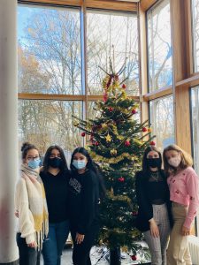 Sechs Personen stehen in einem hellen Raum vor einem festlich geschmückten Weihnachtsbaum. Alle tragen Mund-Nasen-Schutz. Der Baum ist mit goldenen und roten Ornamenten sowie Lichtern dekoriert. Große Fenster im Hintergrund lassen Tageslicht herein.