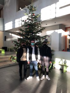 Drei Personen stehen vor einem Weihnachtsbaum in einem hellen Raum. Sie tragen Masken und lächeln. Der Baum ist mit Dekoration geschmückt und es fallen Lichtstrahlen durch ein Fenster auf den Boden. Im Hintergrund sind einige Pflanzen und Stühle sichtbar.