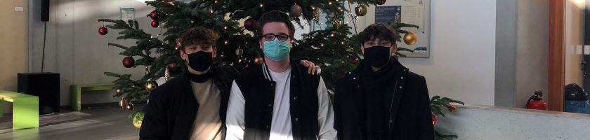 Drei Personen stehen vor einem Weihnachtsbaum in einem hellen Raum. Sie tragen Masken und lächeln. Der Baum ist mit Dekoration geschmückt und es fallen Lichtstrahlen durch ein Fenster auf den Boden. Im Hintergrund sind einige Pflanzen und Stühle sichtbar.