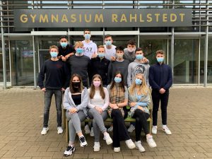 Gruppenfoto von Schülerinnen und Schülern vor dem Gymnasium Rahlstedt. Die Personen tragen Mund-Nasen-Schutzmasken. Sie sitzen und stehen auf einer Bank, lächeln und schauen in die Kamera. Im Hintergrund ist das Schild des Gymnasiums sichtbar.