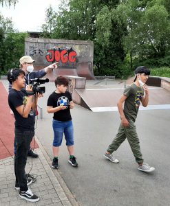 Drei Kinder stehen an einem Skatepark. Ein Junge hält eine Kamera und filmt, während ein anderer Junge ein Smartphone in der Hand hält. Ein drittes Kind geht in Richtung einer Skateboardrampe. Im Hintergrund ist ein Graffiti mit dem Wort 