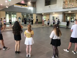 Eine Gruppe von jungen Menschen steht in einem großen Raum, der mit Fenstern und einer hellen Decke ausgestattet ist. Sie sind in einem Kreis angeordnet und scheinen an einer Aktivität oder einem Training teilzunehmen. Die Teilnehmer tragen unterschiedliche Kleidungsstile, darunter Sommerkleider, T-Shirts und Shorts. Im Hintergrund sind weitere Personen sichtbar, die in Gruppen stehen oder sich bewegen. Der Raum hat einen modernen, offenen Charakter.