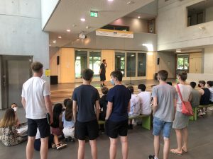 Eine Gruppe von Jugendlichen steht und sitzt in einem modernen Innenraum. Im Vordergrund sind mehrere Personen zu sehen, die in Richtung einer Person blicken, die auf einer kleinen Bühne spricht. Die Umgebung ist hell und freundlich, mit großen Fenstern und minimalistischer Architektur.
