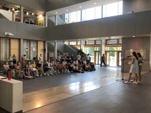 Zwei Personen stehen auf einer kleinen Bühne und sprechen in ein Mikrofon, während eine Gruppe von Menschen, die auf dem Boden sitzt, ihnen zuhört. Im Hintergrund sind große Fenster, die viel Tageslicht hereinlassen, und eine moderne Innenarchitektur mit Treppen und Sitzbereichen ist sichtbar.