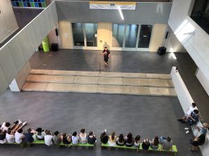 Eine Person steht auf einer kleinen Bühne und spricht zu einer Gruppe von Jugendlichen, die auf einer langen Bank sitzen. Die Szene spielt sich in einem modernen, hellen Raum mit großen Fenstern und minimalistischer Einrichtung ab. Im Hintergrund sind einige Türen und ein Banner sichtbar.