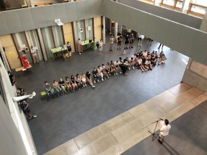 Eine Gruppe von Menschen sitzt in einem großen Raum, während eine Person vorne spricht. Die Anordnung der Sitzplätze ist in mehreren Reihen, und einige Personen stehen. Im Hintergrund sind Tische und Poster sichtbar. Der Raum hat eine moderne Architektur mit großen Fenstern, die viel Tageslicht hereinlassen.