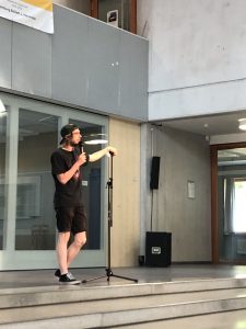 Ein Vortragender steht auf einer Bühne und spricht in ein Mikrofon. Er trägt ein schwarzes T-Shirt und Shorts und hat einen Hut auf. Im Hintergrund ist eine moderne, helle Innenarchitektur mit großen Fenstern und einer offenen Raumgestaltung zu sehen.