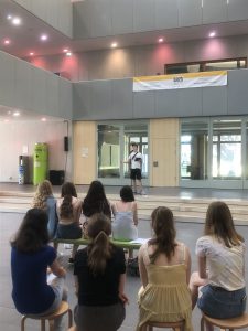 Eine Gruppe von jungen Menschen sitzt auf Hockern und schaut auf eine Bühne, auf der eine Person spricht oder auftritt. Die Umgebung ist hell und modern, mit großen Fenstern und einer Banner über der Bühne.