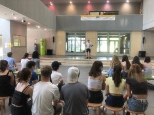 Eine Gruppe junger Menschen sitzt auf Hockern und schaut auf eine Bühne, auf der eine Person spricht. Der Raum ist hell und modern gestaltet, mit großen Fenstern und einem Banner an der Wand. Die Zuhörer sind in lässiger Kleidung und scheinen aufmerksam zuzuhören.