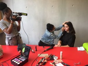 Eine Person filmt mit einer Kamera, während eine andere Person Make-up auf das Gesicht einer sitzenden Person aufträgt. Auf einem roten Tisch liegen verschiedene Make-up-Produkte und Utensilien. Im Hintergrund sind graue Wände sichtbar.