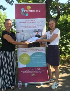 Zwei Frauen stehen nebeneinander und halten ein Dokument in den Händen. Im Hintergrund steht ein Banner mit dem Schriftzug 