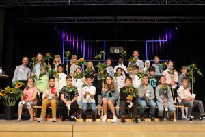 Gruppe von Schülern auf einer Bühne, die fröhlich lächeln und Sonnenblumen in der Hand halten. Die Schüler sind in verschiedenen Kleidungsstilen gekleidet und sitzen auf einer Holzbank. Im Hintergrund sind farbige Lichter und eine Bühne zu sehen.